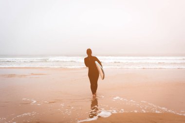 Genç kız garson okyanusun kenarında sörf tahtasıyla duruyor. Fırtınalı hava