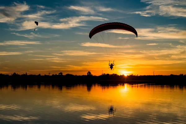 Gün doğumunda gölün üzerinde uçan iki paraşütçü. Suda yansıması olan paramotor pilotu