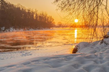 Şafak vakti yumuşak güneş ışığı buzlu nehrin üzerine dökülür, buzlu kıyıları aydınlatır ve sakin bir manzara yaratır.