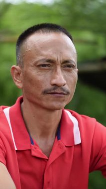 Kırmızı gömlekli düşünceli bir adam, ciddi ifade sakin; yumuşak yeşil arka plan ile açık hava ayarları.