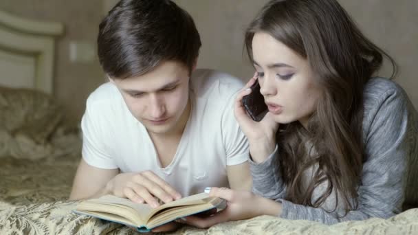 Хлопець читає книжку. Дівчина розмовляє по мобільному телефону. Обидва лежать на ліжку . — стокове відео