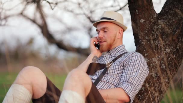 Hombre al aire libre sentado en la hierba hablando en un teléfono inteligente . — Vídeos de Stock