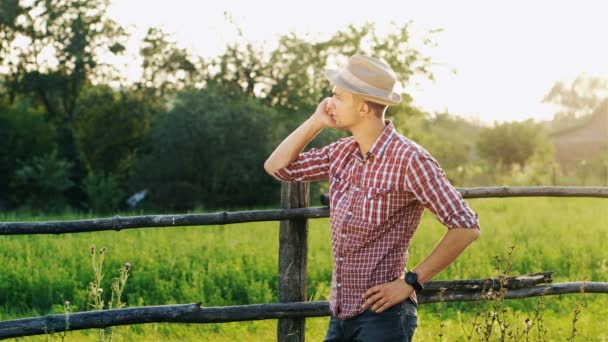 Молодий фермер в капелюсі і перевірена сорочка розмовляє по мобільному телефону на заході сонця . — стокове відео