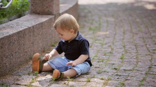 Un niño de un año está sentado en un bloque de piedra en el parque. Día de verano . — Vídeo de stock