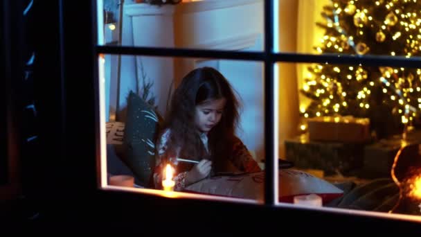 Χαριτωμένο κοριτσάκι βρίσκεται στο κρεβάτι και να γράψει γράμμα στον Άγιο Βασίλη κοντά στο χριστουγεννιάτικο δέντρο και τα δώρα, τα Χριστούγεννα και την Πρωτοχρονιά έννοια — Αρχείο Βίντεο