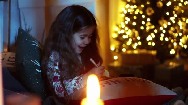 Χαριτωμένο κοριτσάκι βρίσκεται στο κρεβάτι και να γράψει γράμμα στον Άγιο Βασίλη στο δωμάτιο διακοσμημένα για τα Χριστούγεννα, παραμονή Χριστουγέννων — Αρχείο Βίντεο