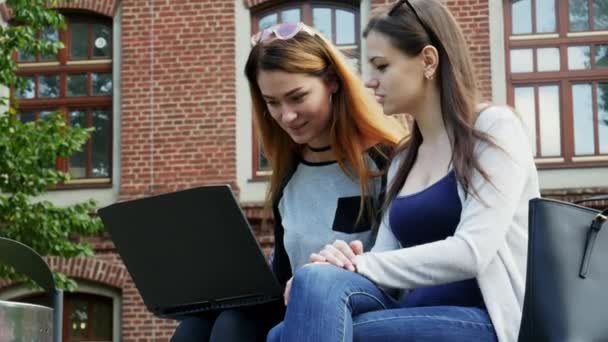 Современные студентки работают вместе над ноутбуком над студенческим проектом, сидя на скамейке в кампусе перед уроками. Образование — стоковое видео