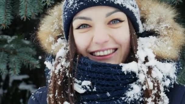 A charming girl with snow on her hair and head looks at the camera in a snowy forest — Stock Video