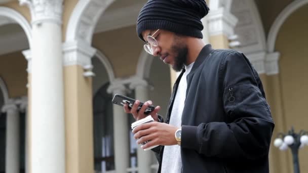 Retrato de hombre de raza mixta, de pie en la calle con una taza de café y el uso de teléfono inteligente — Vídeos de Stock