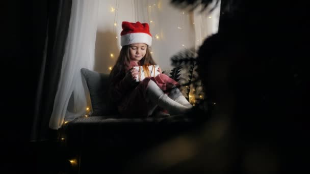 Маленькая девочка в шляпе Санты и рождественском платье сидит на подоконнике дома украшен гирляндой с подарочной коробкой. Новый год и Рождество — стоковое видео