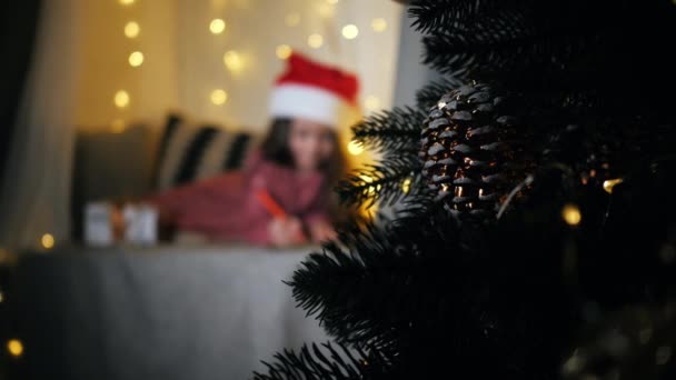 Julgran i fokus i bakgrunden söt barn i Santa hatt skriver ett brev till jultomten på fönsterbrädan, dekorerad för jul, förbereda sig för semester — Stockvideo