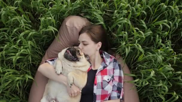 Mujer se acuesta con un pug en el campo en la silla de la bolsa, beso y abrazos perro, jugando en la naturaleza — Vídeos de Stock