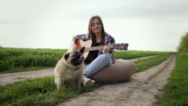Kobieta śpiewa i gra na gitarze z mopsa siedzi na torbie krzesło w zielonym polu — Wideo stockowe