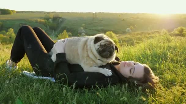 Mujer se acuesta con un pug en la hierba al atardecer, sostiene sus brazos y acaricia al perro — Vídeos de Stock