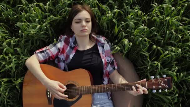 Mujer yace en un campo de trigo toca música en la guitarra y canta, en cámara lenta al atardecer. — Vídeos de Stock