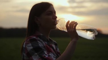 Kız gün batımında susuzluğunu bir tarlada gideriyor. Bir kızın silueti plastik bir şişeden su içer, güneş ışınları 