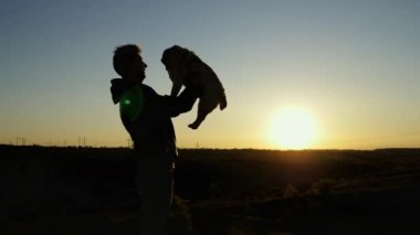 Mutlu bir boksör köpeğini havaya fırlatan adam, gün batımında gökyüzünde sarılıyor.