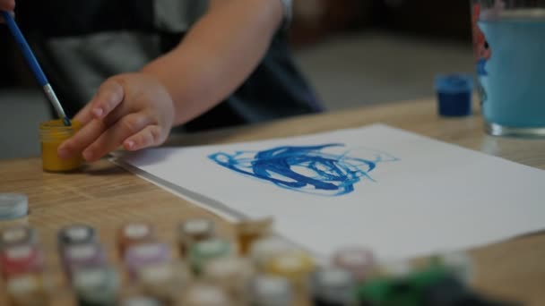 Κοντινό πλάνο των χεριών του μικρού παιδιού αντλεί με ένα πινέλο και μπλε χρώμα σε ένα φύλλο χαρτί στο σπίτι — Αρχείο Βίντεο