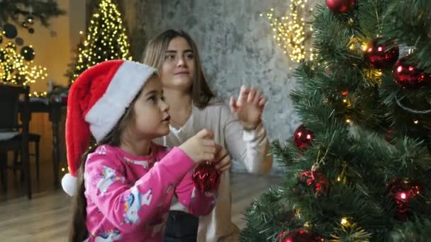 Mutter und Tochter mit Weihnachtsmütze schmücken den Weihnachtsbaum mit Kugeln und bereiten — Stockvideo