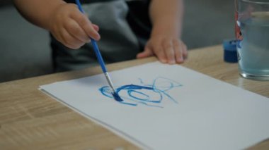 Evdeki bir kağıt parçasına fırça ve mavi boya çizen küçük bir çocuğun ellerine yakın çekim