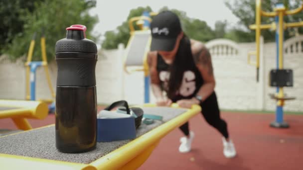 Жінка робить штовхання на спортивному майданчику в розмитому стані, на передньому плані пляшка з водою — стокове відео