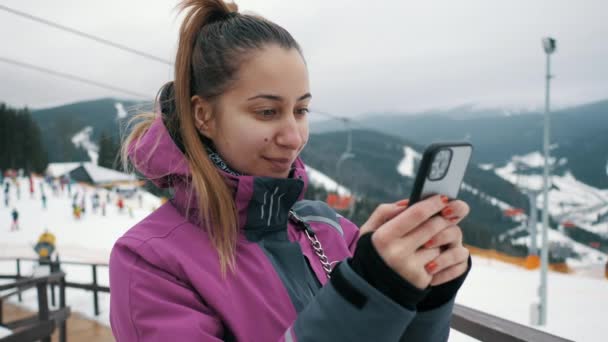 Молода красива жінка використовує мобільний телефон на підйомі гірськолижного курорту, гір і лижників на задньому плані . — стокове відео