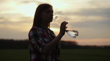 Kadın gün batımında plastik bir şişeden su içiyor. Kadın doğadaki susuzluğunu giderir.