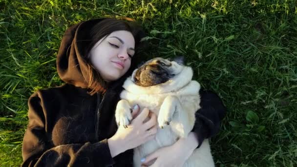 Жінка лежить з мопсом у траві на заході сонця, тримається на руках і кидає собаку — стокове відео