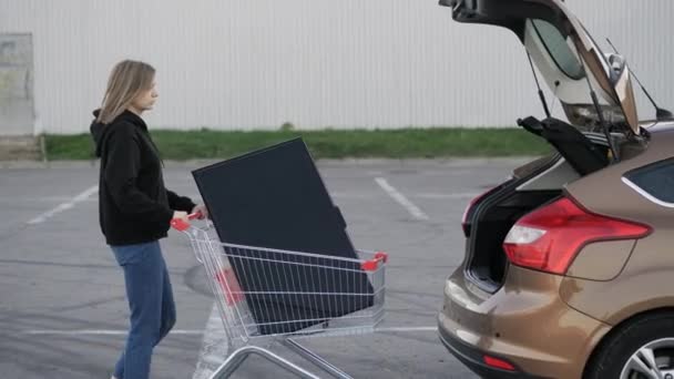 Mujer lleva un televisor grande moderno en un carro de supermercado y trata de cargarlo en el maletero de un coche en el aparcamiento — Vídeos de Stock