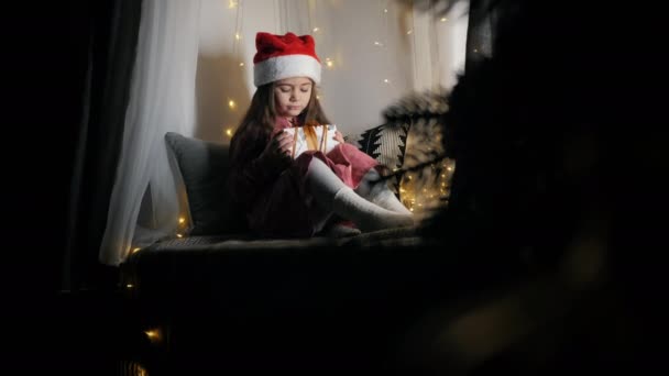 Noel Baba şapkalı küçük tatlı kız, Noel 'i bekliyor, pencere eşiğinde otururken elinde bir hediye tutuyor. — Stok video