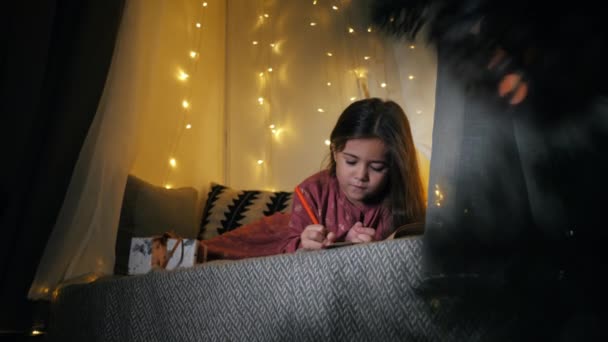 Liten söt flicka skriver ett brev till jultomten med önskningar som ligger på fönsterbrädan dekorerad med en krans, jul — Stockvideo