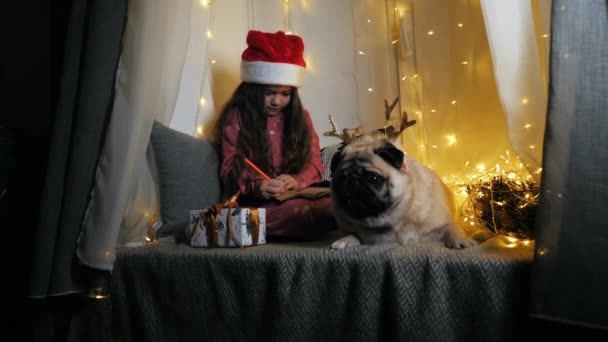 Liten söt flicka med sin roliga mops hund skriver ett brev till jultomten med önskningar sitter på fönsterbrädan — Stockvideo