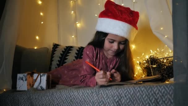 Kleines Mädchen schreibt einen Brief an den Weihnachtsmann mit Wünschen, die auf der Fensterbank liegen und mit einer Girlande geschmückt sind — Stockvideo