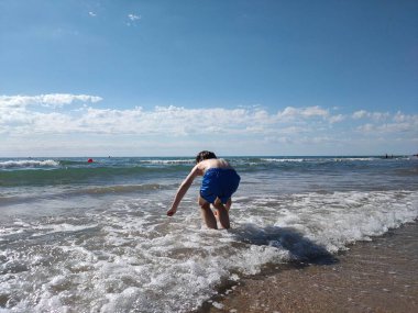 Mavi şortlu çocuk denizde suyla oynuyor.