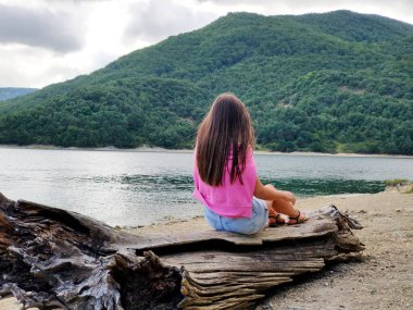 Pembe kazaklı genç bir kız bir dağ gölünün kıyısındaki bir kütüğe oturur.