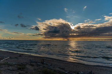 Gün batımı bulutların arkasında su ufkuna yakın sularda parlak güneş ışığının yolunu yansıtıyor ve kumsalın kıyısından her şeyi seyrediyor kumsalda büyük ve küçük ağaçları duruluyor.