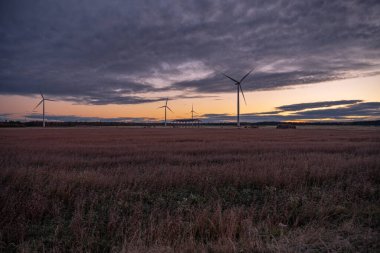 Rüzgar jeneratörleri, saman silindirleri ve çayırdaki bir binanın iskeleti. Rüzgar jeneratörleri orman yakınlarında dönüyor ve elektrik üretiyor. Gökyüzünde bulutlar var.