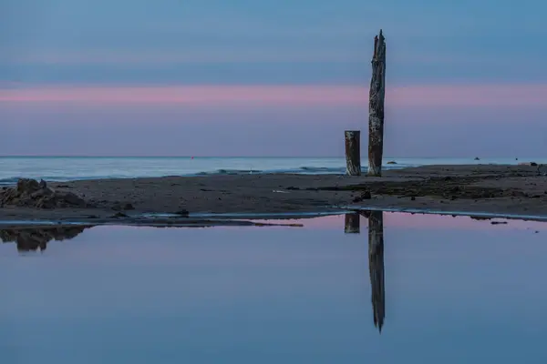 日没後の海のそばの古い木の柱と隣の小さな水槽のポールの反射 — ストック写真