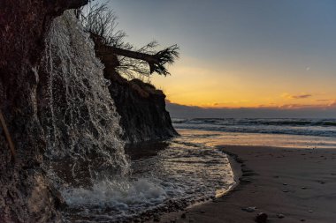 Gün batımı şelalesi deniz kıyısında. Geniş deniz sularında küçük güzel bir nehir yatağı ve ufukta bulutlar var.