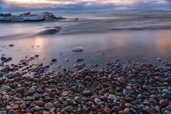 海水の表面を照らし空の色のついた石や雲を照らす夕日のある海岸の小さな石 — ストック写真