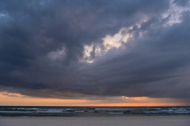 Gün batımında deniz kenarında kara bulutlar küçük dalgalar ve güneş bulutların arkasına dağılmış sadece ufukta turuncu bir çizgi bırakıyor.