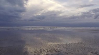 Okyanus dalgaları sahili turluyor. Kuma yansıyan gökyüzü bulutları.