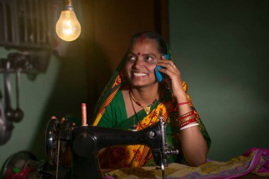 Hintli kırsal bir kadın dikiş makinesi kullanıyor ve cep telefonuyla konuşuyor.