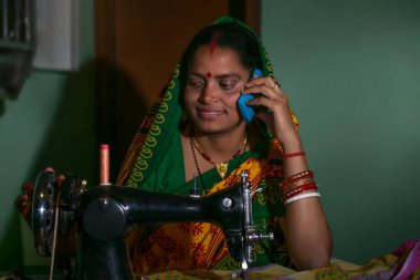 Hintli kırsal bir kadın dikiş makinesi kullanıyor ve cep telefonuyla konuşuyor.