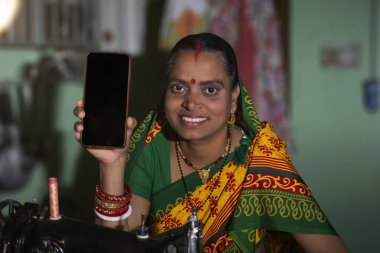 Hintli kırsal bir kadın dikiş makinesi kullanıyor ve mobil telefonunu gösteriyor.