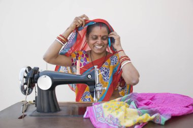 Hintli kırsal bir kadın dikiş makinesi kullanıyor ve cep telefonuyla konuşuyor.