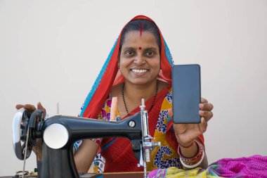 Hintli kırsal bir kadın dikiş makinesi kullanıyor ve mobil telefonunu gösteriyor.