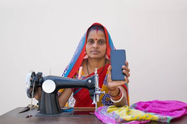 Hintli kırsal bir kadın dikiş makinesi kullanıyor ve mobil telefonunu gösteriyor.