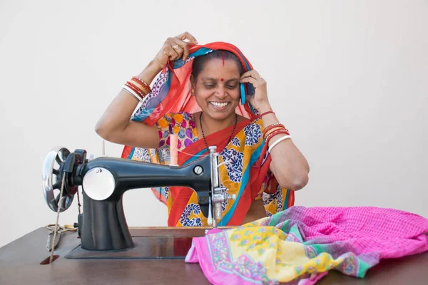 Hintli kırsal bir kadın dikiş makinesi kullanıyor ve cep telefonuyla konuşuyor.