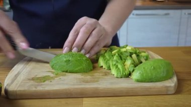 Kadın şef, mutfak bıçağıyla ahşap bir masada avokadoyu küçük küplere ayırıyor.